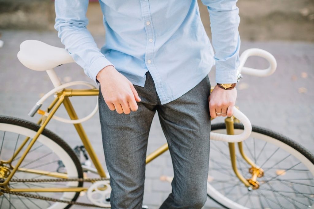 A man in a light blue shirt and gray pants stands next to a stylish gold bicycle, holding its handlebars.