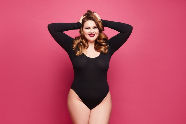 Young plus-size model with bright makeup wearing black bodysuit on pink background.
