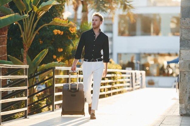 Man walking outdoors with luggage, dressed in white pants and black shirt, stylish travel look in sunny urban setting.