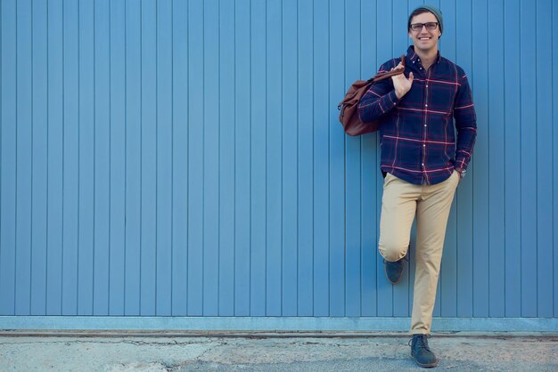 Man in a navy checkered shirt and beige pants leaning against a blue wall, carrying a brown bag and smiling confidently.