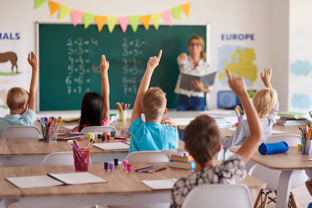 Young students raise hands eagerly while a teacher stands at the front near a chalkboard in a bright classroom. 